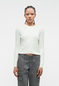 Biely cardigan s pleteným vzorom, dlhé rukávy a skrátený strih, s okrúhlym výstrihom a piatimi gombíkmi vpredu. Šedé cargo nohavice.