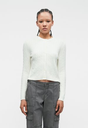 Witte kabelgebreide cardigan met lange mouwen en een cropped pasvorm, voorzien van een ronde halslijn en vijf knopen aan de voorkant. Grijze cargobroek.