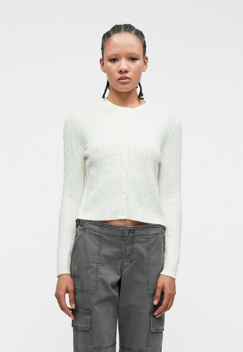 Biely cardigan s pleteným vzorom, dlhé rukávy a skrátený strih, s okrúhlym výstrihom a piatimi gomb�íkmi vpredu. Šedé cargo nohavice.