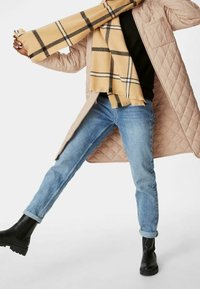 Beige vadderad kappa, svart tröja, ljusblå jeans och svarta ankelskor. Gulrutigt scarf med svarta detaljer hängande över axlarna.