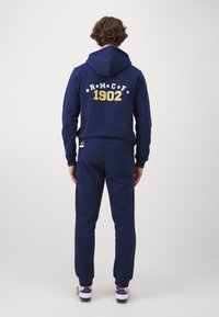 Sweatshirt azul-marinho com bolso canguru, apresentando texto branco e dourado "R M C F 1902" nas costas. Combinado com calças de fato à condizer.