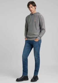Sudadera gris de punto con capucha ajustable con cordón, combinada con vaqueros azules de denim. El modelo lleva botines negros. Diseño sencillo con acentos mínimos.