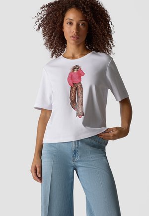 Woman with curly hair wears a white t-shirt with a graphic of a woman in a pink sweater and patterned wide-leg pants, paired with light blue jeans.