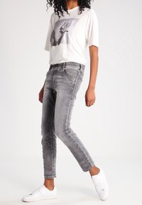 Szare dopasowane dżinsy z wyblakłą fakturą, zestawione z luźną kremową koszulką z czarno-białą grafiką oraz białymi butami wsuwanymi.