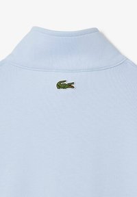Polo di colore azzurro chiaro con colletto ripiegato e logo di coccodrillo ricamato in verde sul retro. Tessuto morbido con una texture liscia.
