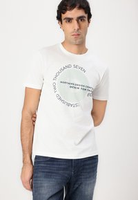 Hvit bomulls-T-skjorte med et rundt trykk med tekst i blått og grønt, rund halslinning og korte ermer, sammen med blå jeans.