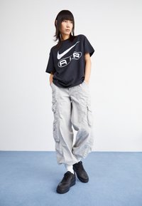 Henkilö, jolla on yllään musta Nike Air -t-paita, vaaleanharmaat cargo-housut ja mustat lenkkarit, seisoo sinisellä lattialla valkoista seinää vasten.