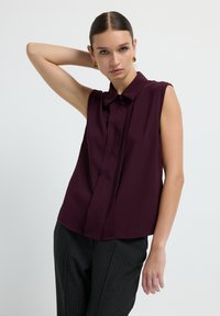 Blusa bordeaux a maniche corte con colletto a bottoni, sottili pieghe frontali e una consistenza leggera e liscia. Abbinata a pantaloni grigi a righe.