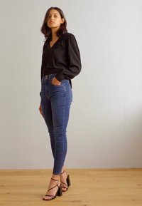 Blusa nera di satin con maniche lunghe, abbinata a jeans blu attillati e sandali neri con tacco a stiletto su un pavimento di legno.