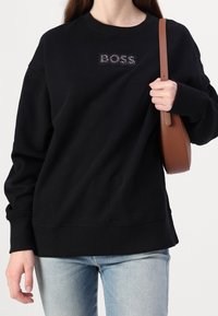 Svart sweatshirt med ribbade muddar och nederkant, med en texturerad "BOSS"-logotyp. Tillsammans med ljusblå denim och en brun crossover-väska.