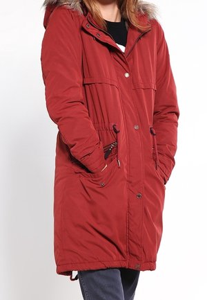 Personne portant un long manteau d'hiver rouge avec une capuche bordée de fourrure et les mains dans les poches avant sur un fond uni.