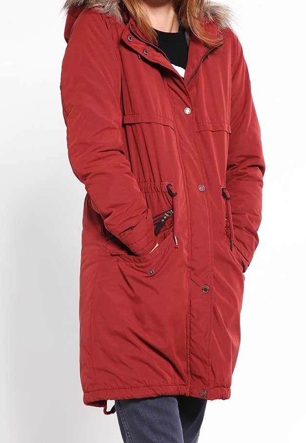 Personne portant un long manteau d'hiver rouge avec une capuche bordée de fourrure et les mains dans les poches avant sur un fond uni.