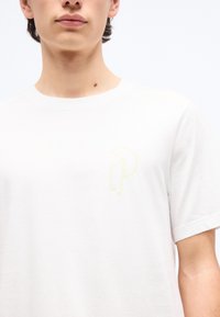 Νεαρό άτομο που φοράει απλή λευκή κοντομάνικη μπλούζα με διακριτικό κίτρινο γεωμετρικό λογότυπο στο αριστερό στήθος, με φόντο ένα λευκό τοπίο.