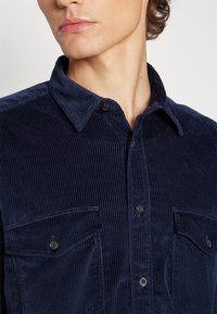 Jeune personne portant une chemise en velours côtelé bleu foncé avec des poches poitrine boutonnées, légèrement tournée vers la gauche sur un fond uni.
