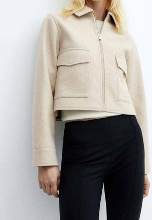Femme portant une veste courte zippée beige avec de grandes poches à rabat et un pantalon noir ajusté sur fond blanc.