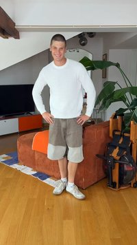 Νέος άνδρας με λευκό πουλόβερ και μπεζ σορτς στέκεται γελώντας σε ένα σαλόνι με πορτοκαλί καναπέ και εσωτερικό φυτό.