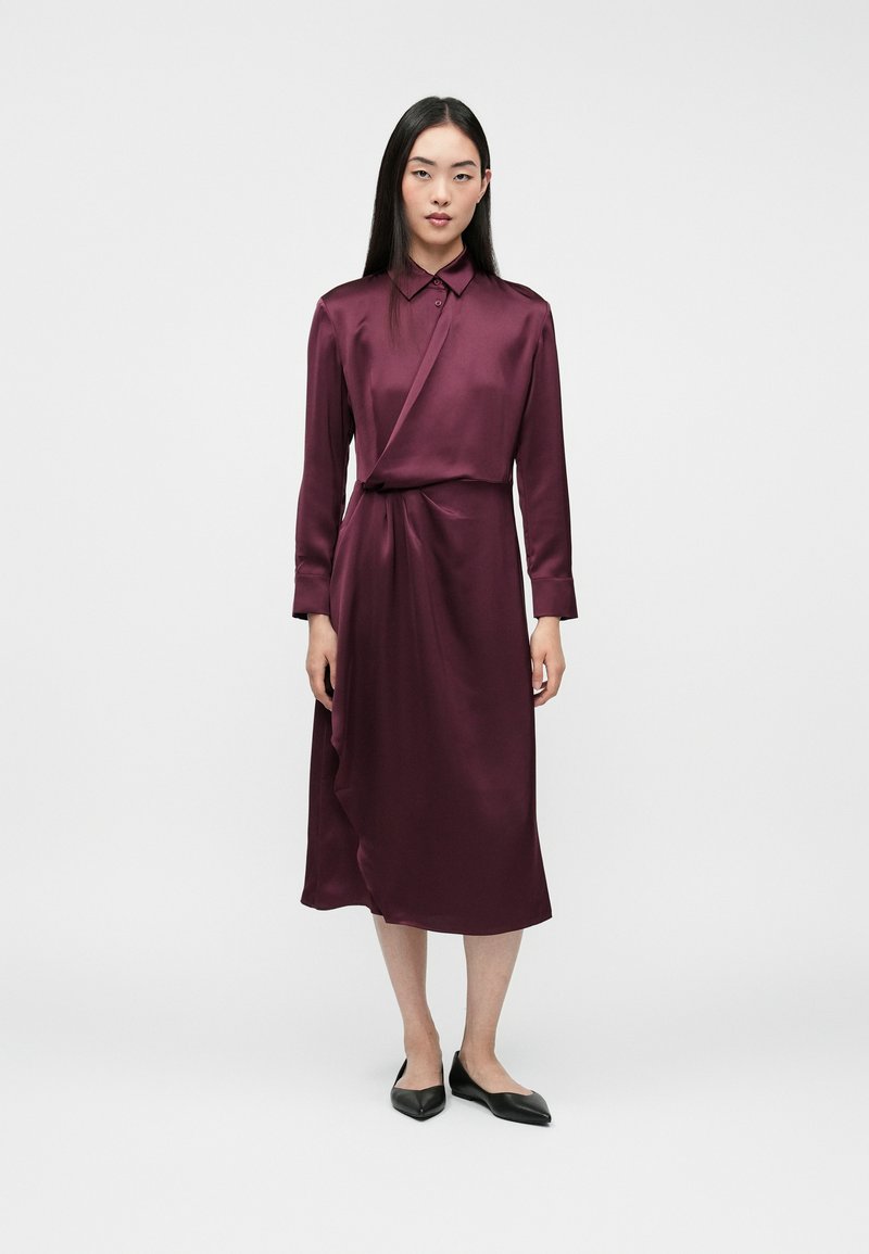 Bordowa sukienka z satyny z kołnierzykiem, długimi rękawami i krojem kopertowym, z bocznym węzłem. Noszona z czarnymi szpiczastymi balerinami, na białym tle.