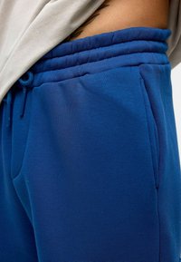 Blauwe sweatpants met een trekkoord in de taille, zijzakken en een zachte, gladde textuur. De stof lijkt lichtgewicht en casual.
