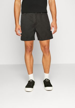 Shorts en coton noir à coupe classique, avec passants de ceinture et finitions ourlées. Portés avec des baskets noires et des chaussettes blanches.