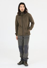 Olijfgroene fleece jas met een hoge kraag, met ritszakken; gecombineerd met grijze broek met donkerdere kniestukken en ritszakken aan de zijkant.