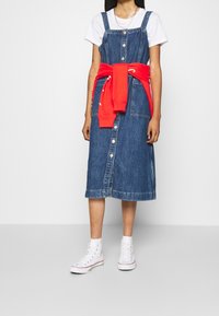 Sukienka ogrodniczka z denimu o długości midi, z guzikami z przodu, regulowanymi ramiączkami i czerwoną bluzą związana w talii. Białe sneakersy.