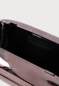 Metaliczna fioletowa kopertówka o smukłym, prostokątnym kształcie, z łańcuszkiem i czarnym wnętrzem z etykietą marki.