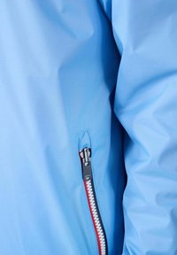 Hellblaue wasserdichte Jacke mit Reißverschlusstasche, kontrastierendem roten Reißverschlussband und glatter Textur. Geeignet für den Außenbereich.
