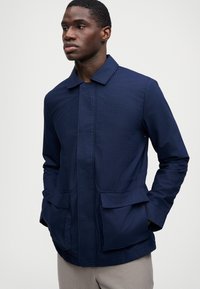 Homem a usar um casaco xadrez azul-escuro com bolsos de aba e calças cinzas claras, de pé com as mãos nos bolsos, olhando para a esquerda.