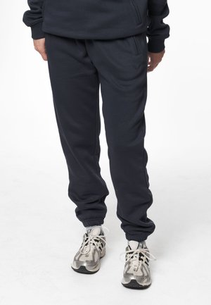 Personne portant un pantalon de jogging noir et des chaussures de course blanches et grises, debout sur un fond blanc.