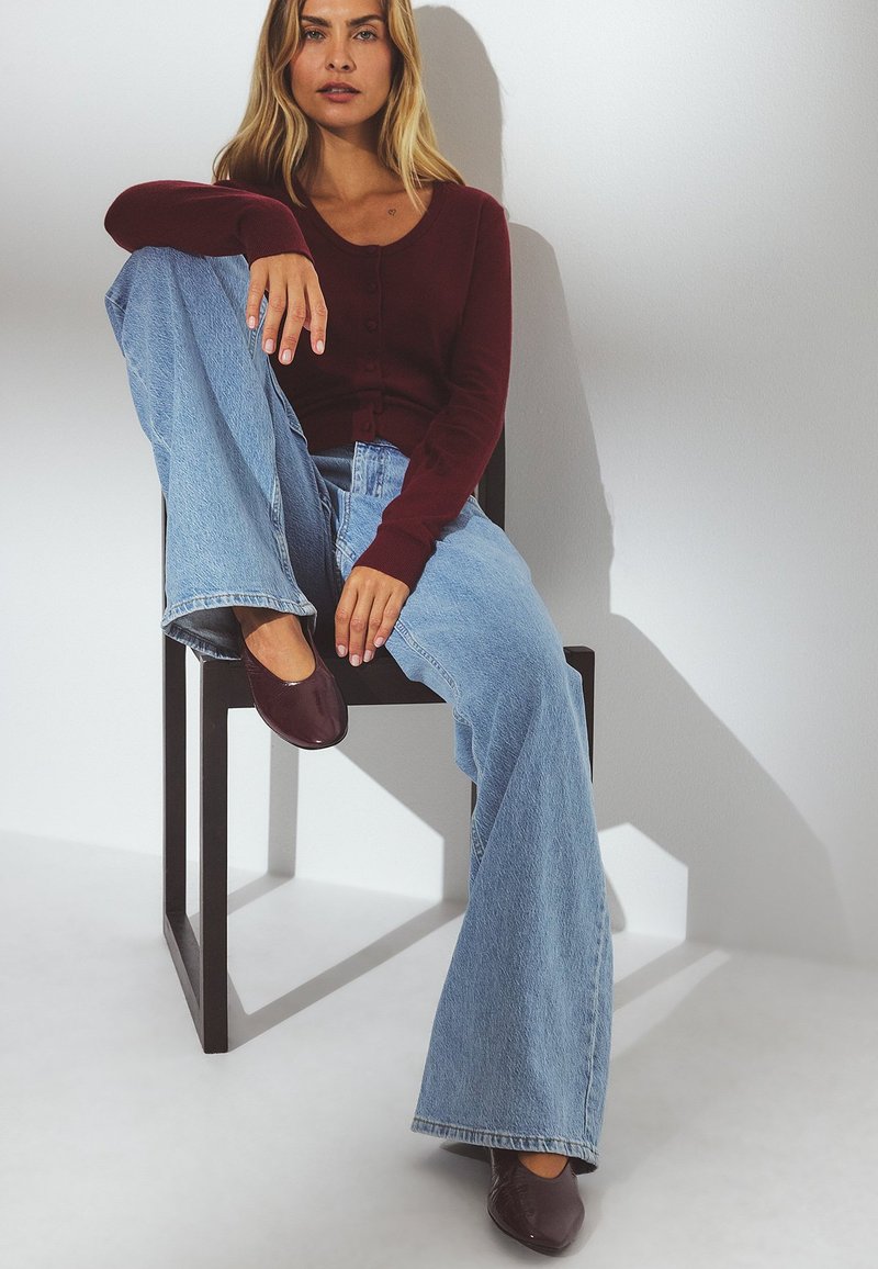 Cardigan bordeaux à boutons, jeans bleu clair à jambes larges et ballerines bordeaux foncé. Modèle assis sur une chaise noire devant un fond neutre.