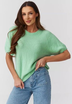Kobieta o długich, brązowych włosach, ubrana w miętowozielony sweter z krótkim rękawem i niebieskie jeansy, pozująca z jedną ręką w kieszeni i delikatnym uśmiechem.
