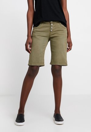 Shorts en denim vert olive avec une coupe droite, fermeture à cinq boutons et poches latérales. Portés avec un haut noir à manches courtes et des chaussures noires à enfiler.