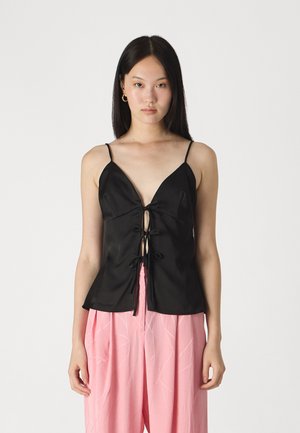 Haut sans manches en satin noir avec décolleté en V profond et fermeture à cordon sur le devant, associé à un pantalon rose pâle à jambes larges avec des motifs subtils.
