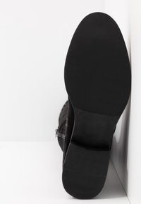 Semelle de botte noire vue de face avec la taille 37 estampillée, reposant verticalement contre un coin blanc en arrière-plan.