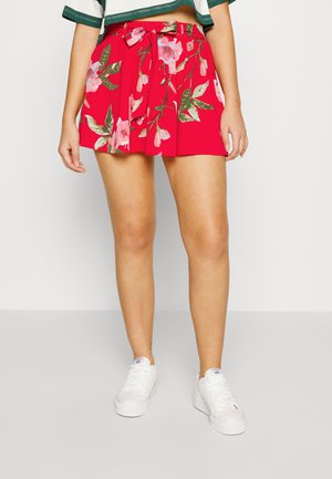 Skort floral vermelho com um cinto laço, design plissado e padrões de folhas verdes. Usado com ténis brancos para um look casual.