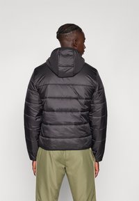 Black puffer jacket with a hood, featuring a quilted design and elastic cuffs. Worn over olive green pants, showing a streamlined silhouette.