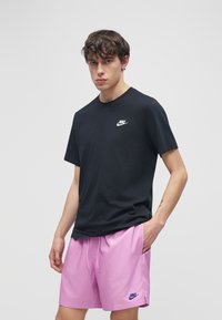 Νέος άνδρας που φοράει μαύρο t-shirt Nike και ροζ σορτς Nike, στέκεται με το ένα χέρι στην τσέπη σε απλό φωτεινό φόντο.