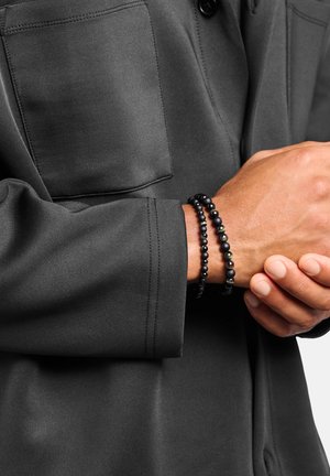 Schwarze Perlenarmbänder, eines mit matten und glänzenden Perlen, die an einem Handgelenk getragen werden. Die Person trägt ein dunkelgraues Hemd mit einer geknöpften Tasche.