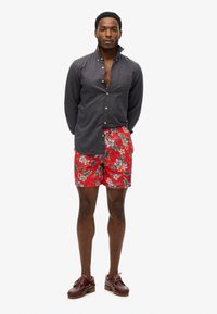 Camisa de botones en gris oscuro, combinada con shorts de baño rojos con flores y hojas verdes. Zapatos marrones con cordones y suelas de goma.