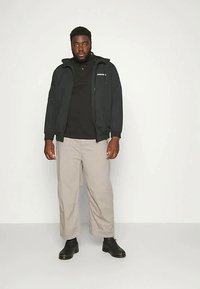 Egy férfi áll egy fehér falnak dőlve, fekete dzsekit, fekete inget, bézs nadrágot és fekete csizmát visel, és közvetlenül előre néz.