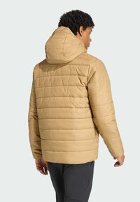 En person med krøllet hår har en beige pufferjakke med hætte på, set fra ryggen, mod en lys grå baggrund.