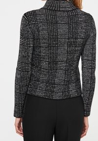 Pull à carreaux noirs et blancs avec un col montant, présentant un motif en maille texturée et une coupe ajustée, associé à un pantalon noir.
