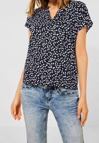 Kvinna iförd en marinblå kortärmad topp med vita prickar och ljusblå urtvättade jeans, ståendes mot en enfärgad bakgrund.