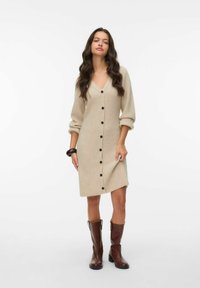 Beige ribbad stickad klänning med V-ringning, lång ärm och svarta knappar fram. Bärs med bruna knähöga stövlar.