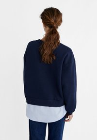 Felpa oversized blu scuro con orlo e polsini a coste, indossata sopra una camicia a righe leggera blu e bianca, crea un look informale.