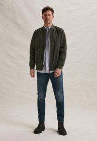 Mann steht und trägt eine grüne Bomberjacke, ein gestreiftes Hemd, blaue Jeans und schwarze Schnürstiefel vor einem schlichten hellen Hintergrund.