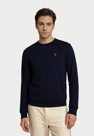 Junger Mann trägt einen dunkelblauen Sweatshirt mit rotem Logo und hellbeige Hose und steht vor einem schlichten weißen Hintergrund.