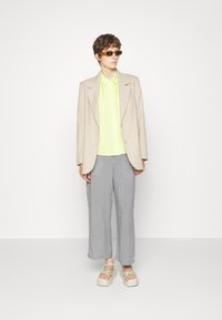 Blazer bege, camisa de colarinho verde neon, calças cinzas com padrão e sandálias de plataforma grossas com tiras verdes; modelo a usar óculos de sol.