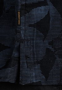 Tejido negro con un patrón gráfico en azul oscuro que presenta rayas horizontales y formas de hojas. Pequeña etiqueta de marca con "PME LEGEND" visible.