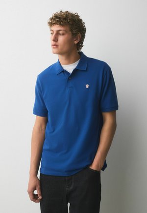 Fiatal férfi göndör hajjal, kék polóingben és fekete nadrágban, egy kézzel a zsebében áll, oldalra néz, egyszínű háttér előtt.